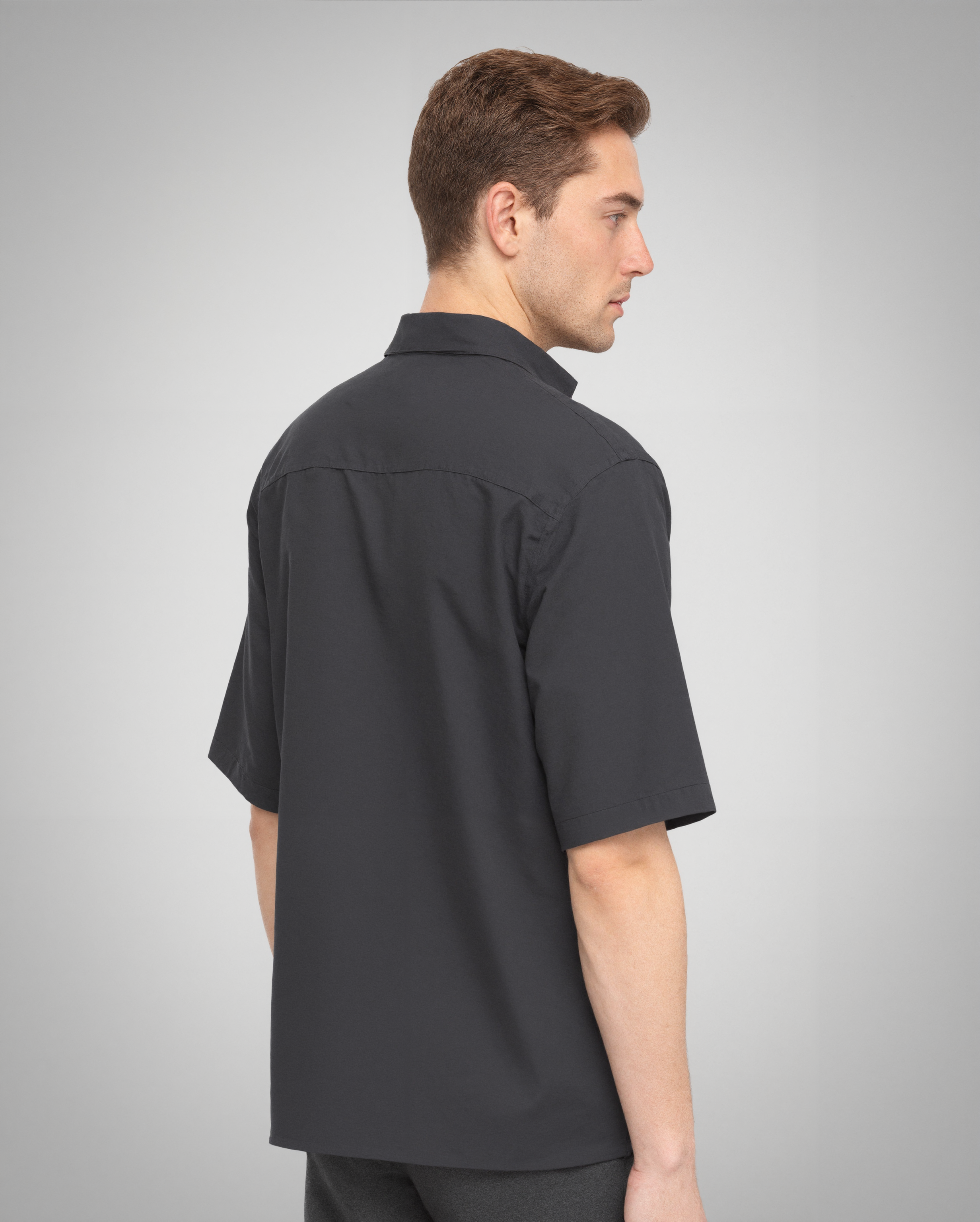 A rear view of a male model wearing a black short-sleeve button-up shirt with a regular fit. The clean back panel showcases the smooth texture of the 100% organically grown cotton fabric. The double-stitched hem and structured collar add subtle detailing to the minimalist design.