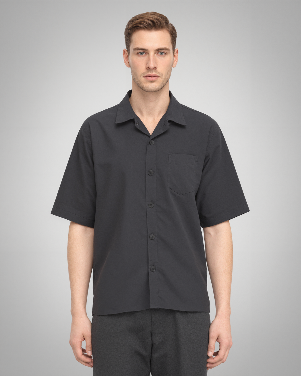 A male model wearing a black short-sleeve button-up shirt with a regular fit, crafted from 100% organically grown cotton. The shirt features a structured spread collar, a single square chest pocket, and a five-button front closure. Styled with dark trousers for a modern, minimalist look.