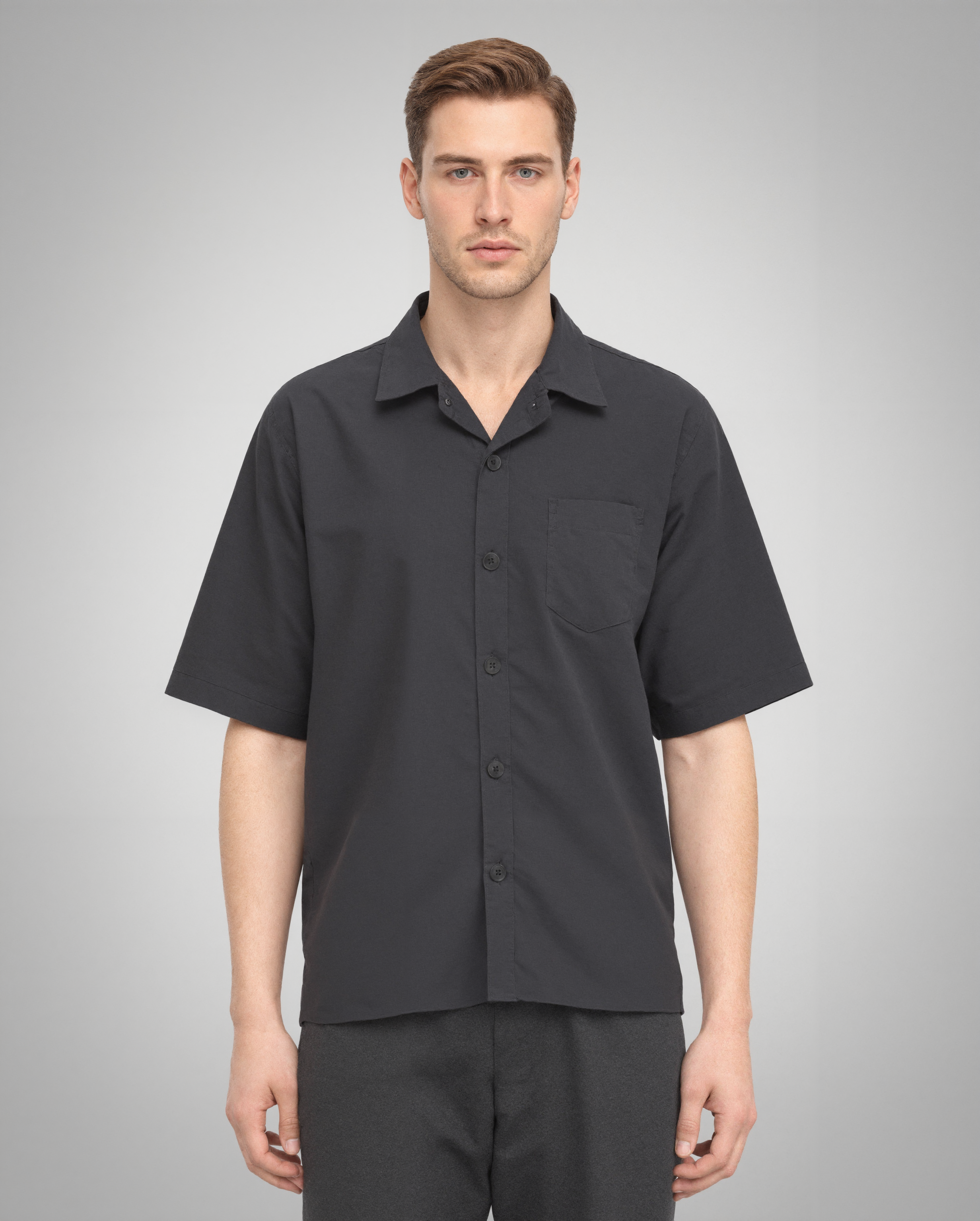 A male model wearing a black short-sleeve button-up shirt with a regular fit, crafted from 100% organically grown cotton. The shirt features a structured spread collar, a single square chest pocket, and a five-button front closure. Styled with dark trousers for a modern, minimalist look.