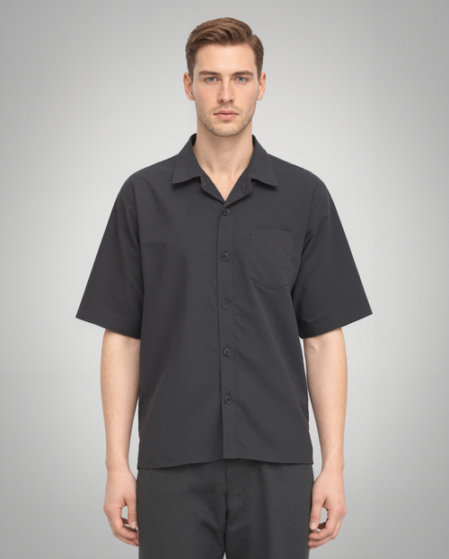 A male model wearing a black short-sleeve button-up shirt with a regular fit, crafted from 100% organically grown cotton. The shirt features a structured spread collar, a single square chest pocket, and a five-button front closure. Styled with dark trousers for a modern, minimalist look.