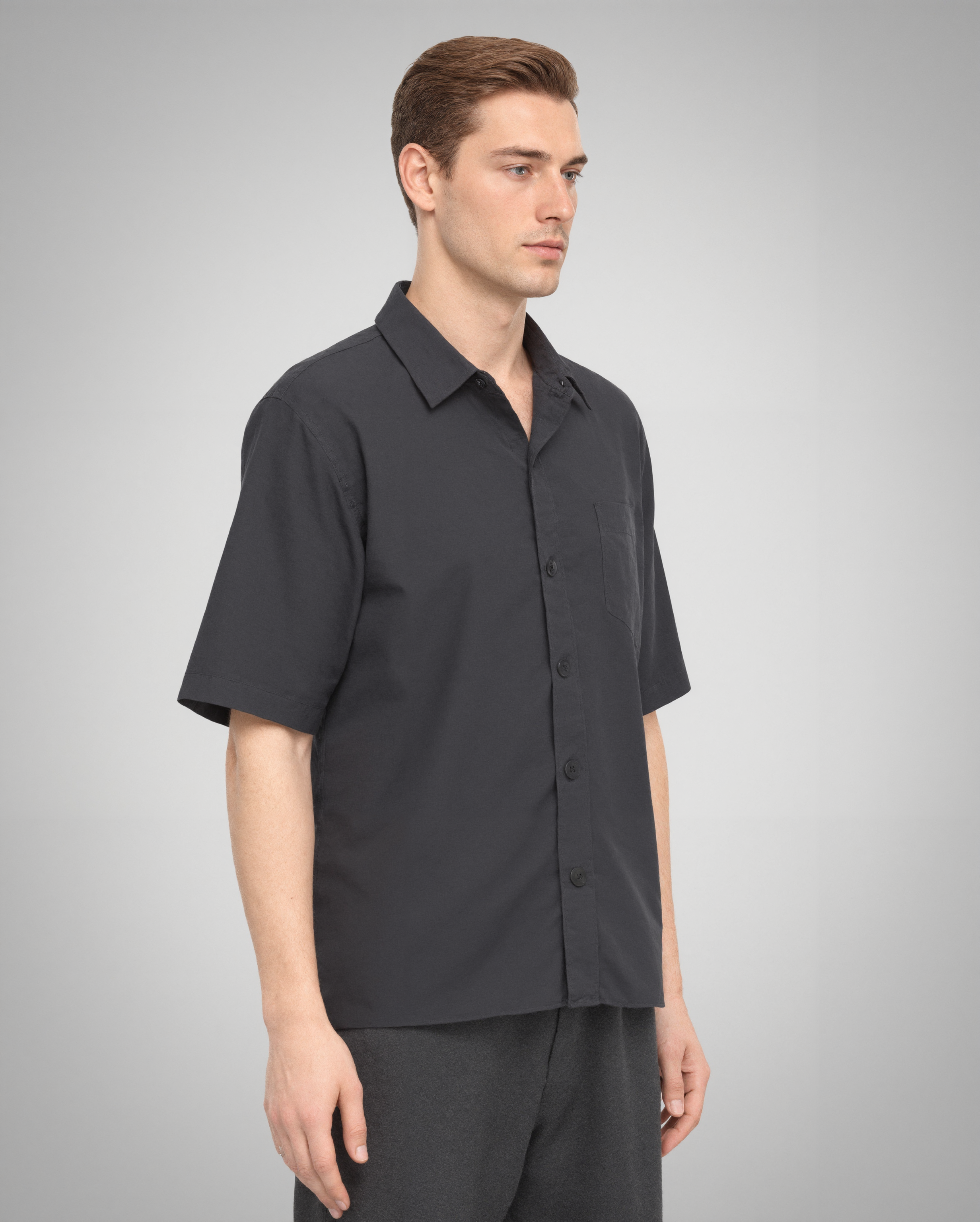A side profile of a male model wearing a black short-sleeve button-up shirt with a regular fit. The organic cotton fabric drapes naturally, and the square chest pocket with topstitching is visible. The structured collar maintains its shape, enhancing the refined aesthetic.